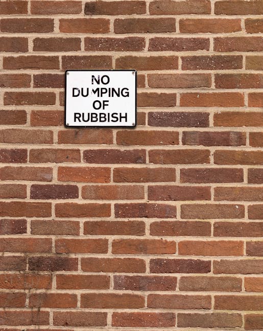 A rectangular white metal sign with black lettering reading 'NO DUMPING OF RUBBISH' is mounted on a red and brown brick wall. The bricks are arranged in a horizontal pattern with light gray mortar bet